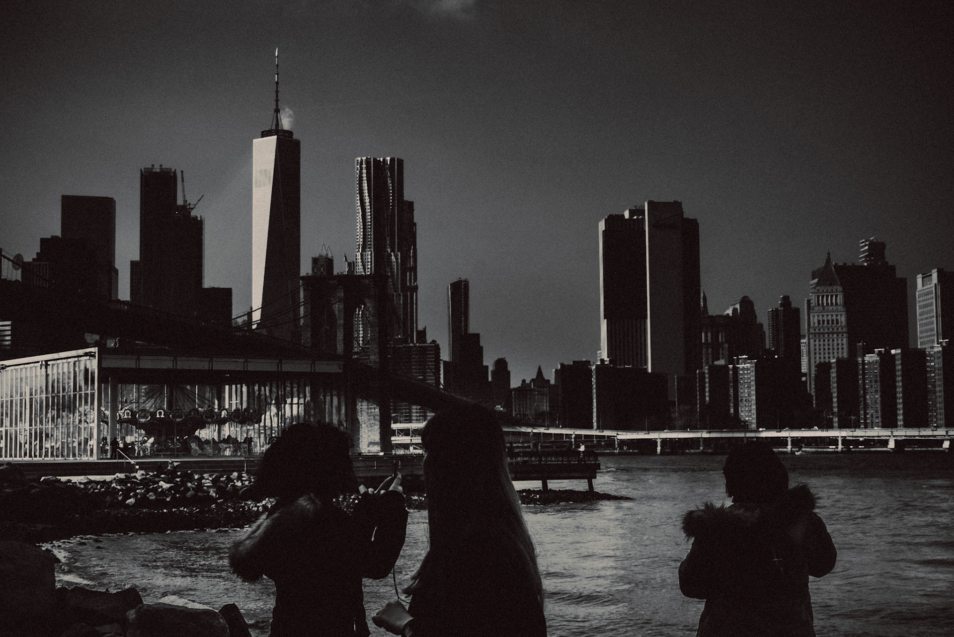 A black and white photo of the Manhattan skyline, One World Trade Center, the Brooklyn Bridge and Jane's Carousel from Pebble Beach in Dumbo, Brooklyn, New York City, USA, December 2017, Sony A7S II.
