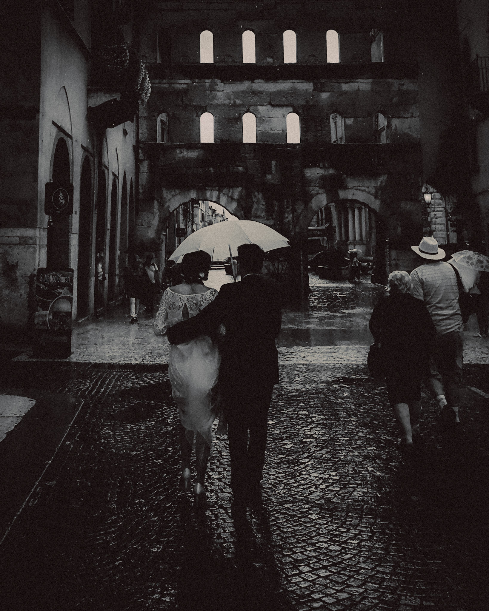 A black and white candid photo of a newly married couple sharing an umbrella under the rain, Verona, Italy, September 2017, Huawei Mate 9 Pro.