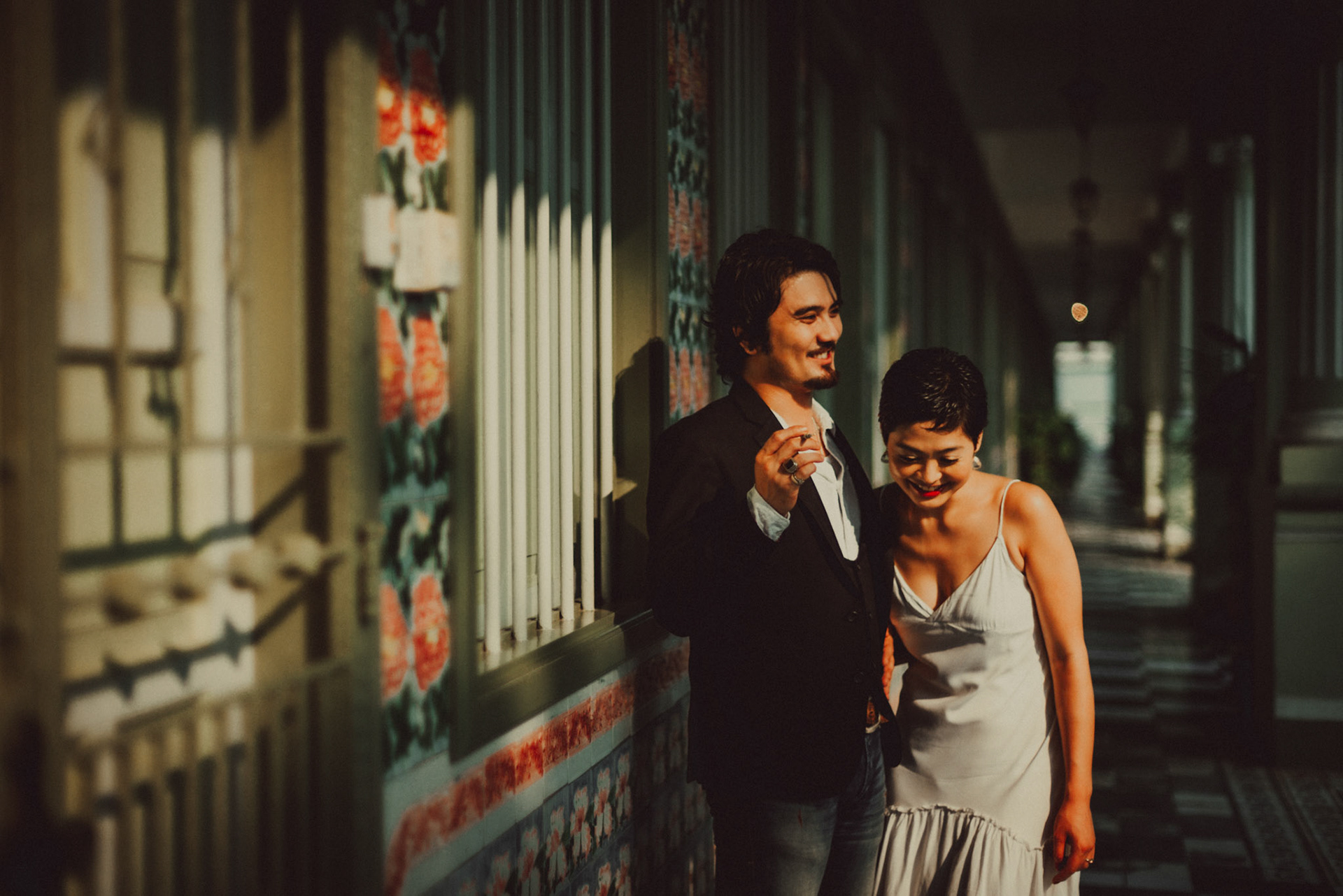 A Japanese couple sharing a lighthearted moment in a sunlit corridor with Peranakan details, from Ibuki and Emi's candid chill engagement shoot, Pertain Road, Little India, Singapore, October 2015, Leica M.