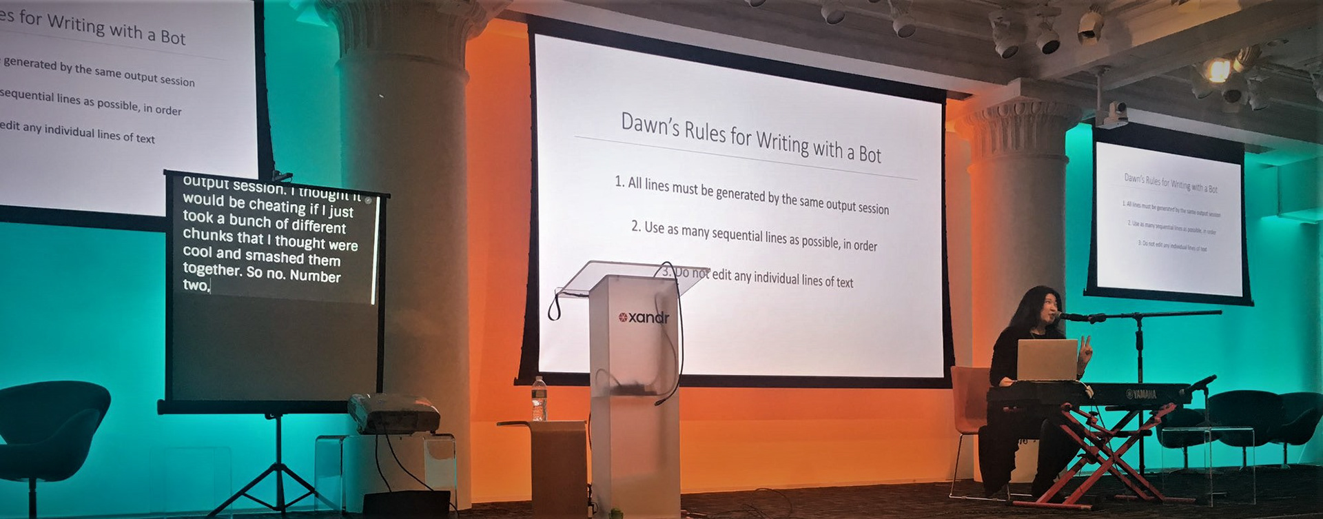 Dawn onstage at !!Con in NYC. She is sitting at a keyboard (instrument) with a large screen behind her that reads: "Dawn's Rules for Writing with a Bot."