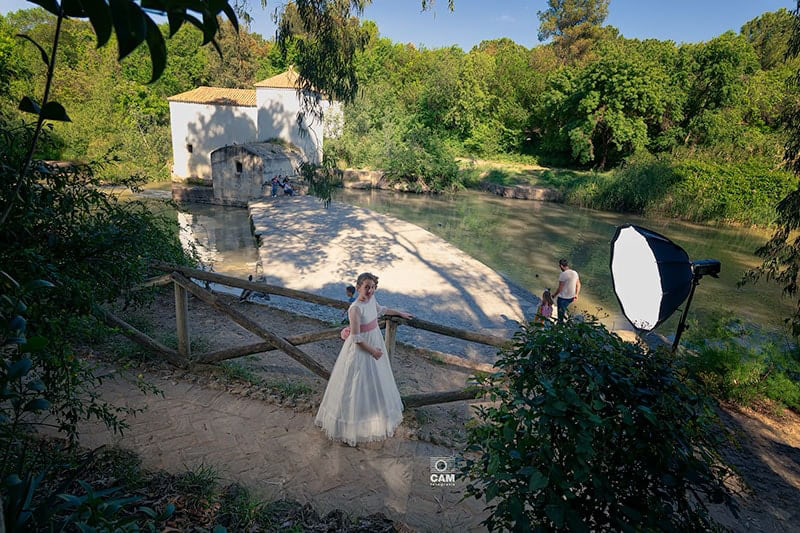 Niña vestida de comunión, con traje blanco en camino de un parque, donde tiene en frente un Softbox de luz de estudio en frente, ella mira a su derecha a la cámara, de fondo se observa un lago y un molino, con algunas personas cruzando el puente sobre el lago.