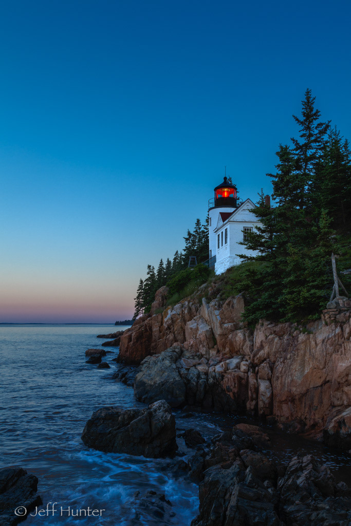 A lighthouse with a red light on a rugged cliff overlooking the ocean at sunset