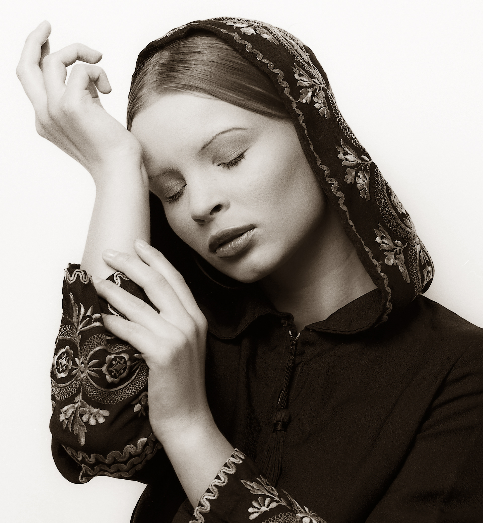 Caucasian woman, eyes closed with ornamental hoodie with posing hands against a white background. Photographed in black and white