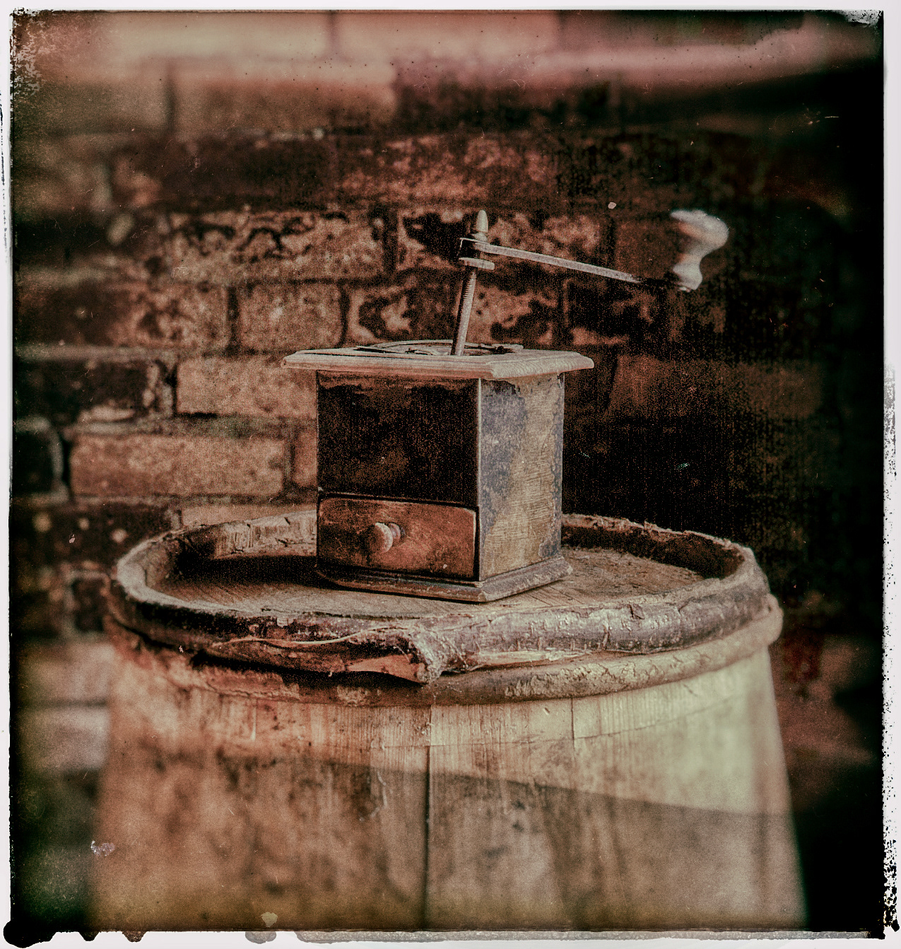 An old coffee grinder on an old wooden barrel against a brick wall. Photographed with a distressed smudgy look
