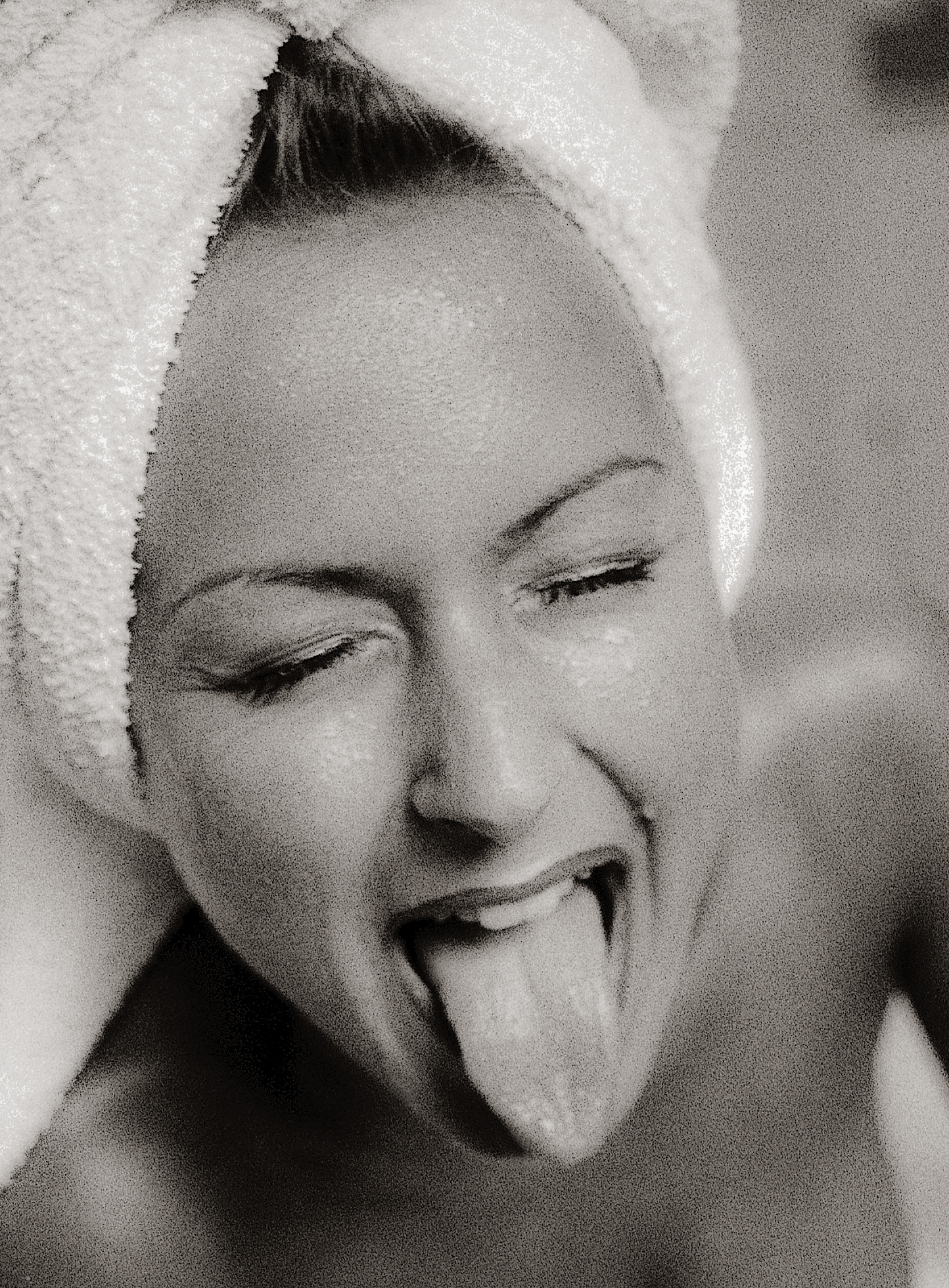 Close up of caucasian womans face, eyes closed, with her tongue out, towel wrapped around her head and water drops on her skin. Photographed in black and white.