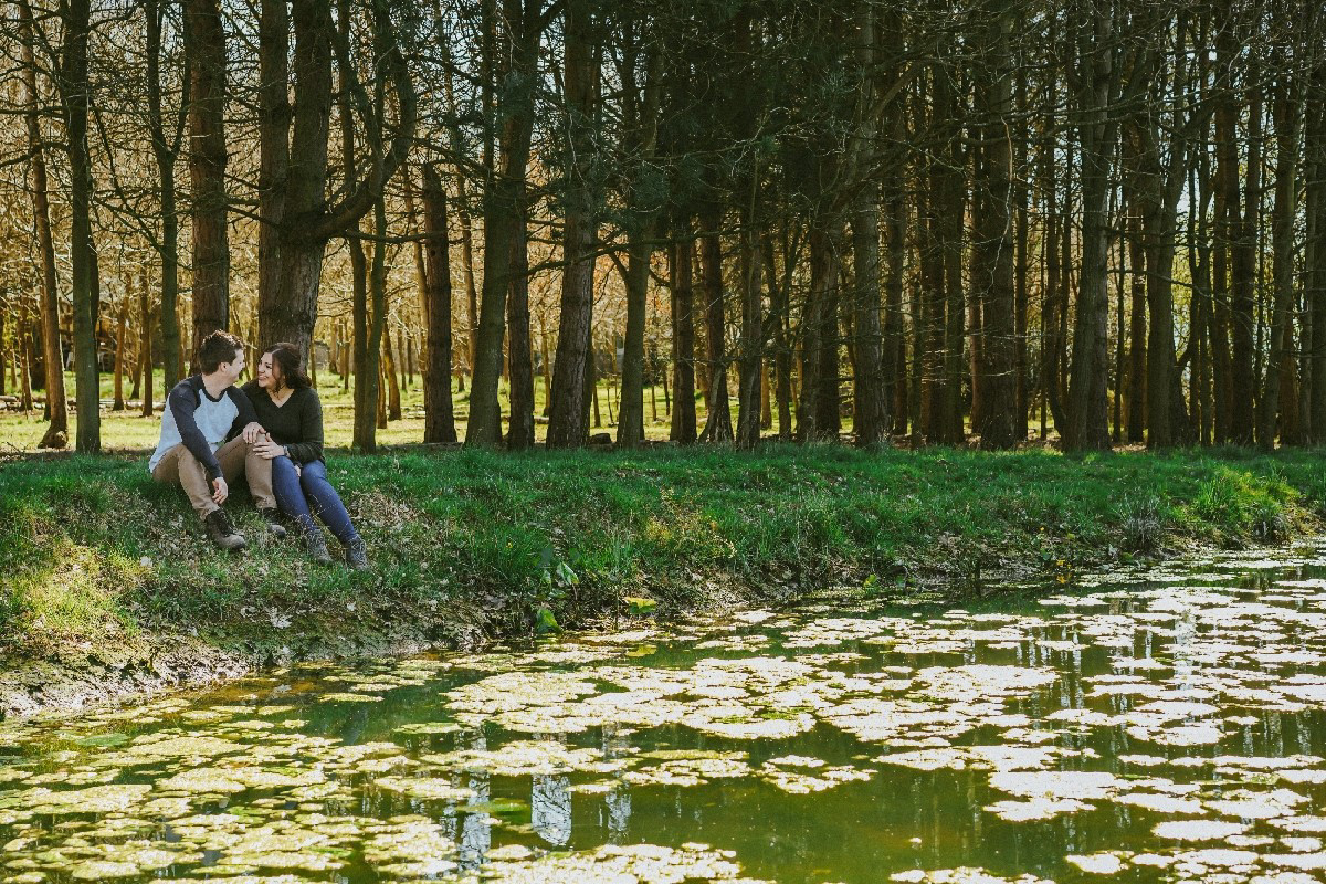 smiling soon to be bride and groom sat on bank of lake at endeavour woodland wedding venue in buckinghamshire during pre wedding engagement photo session with david lefebvre a photographer from milton keynes