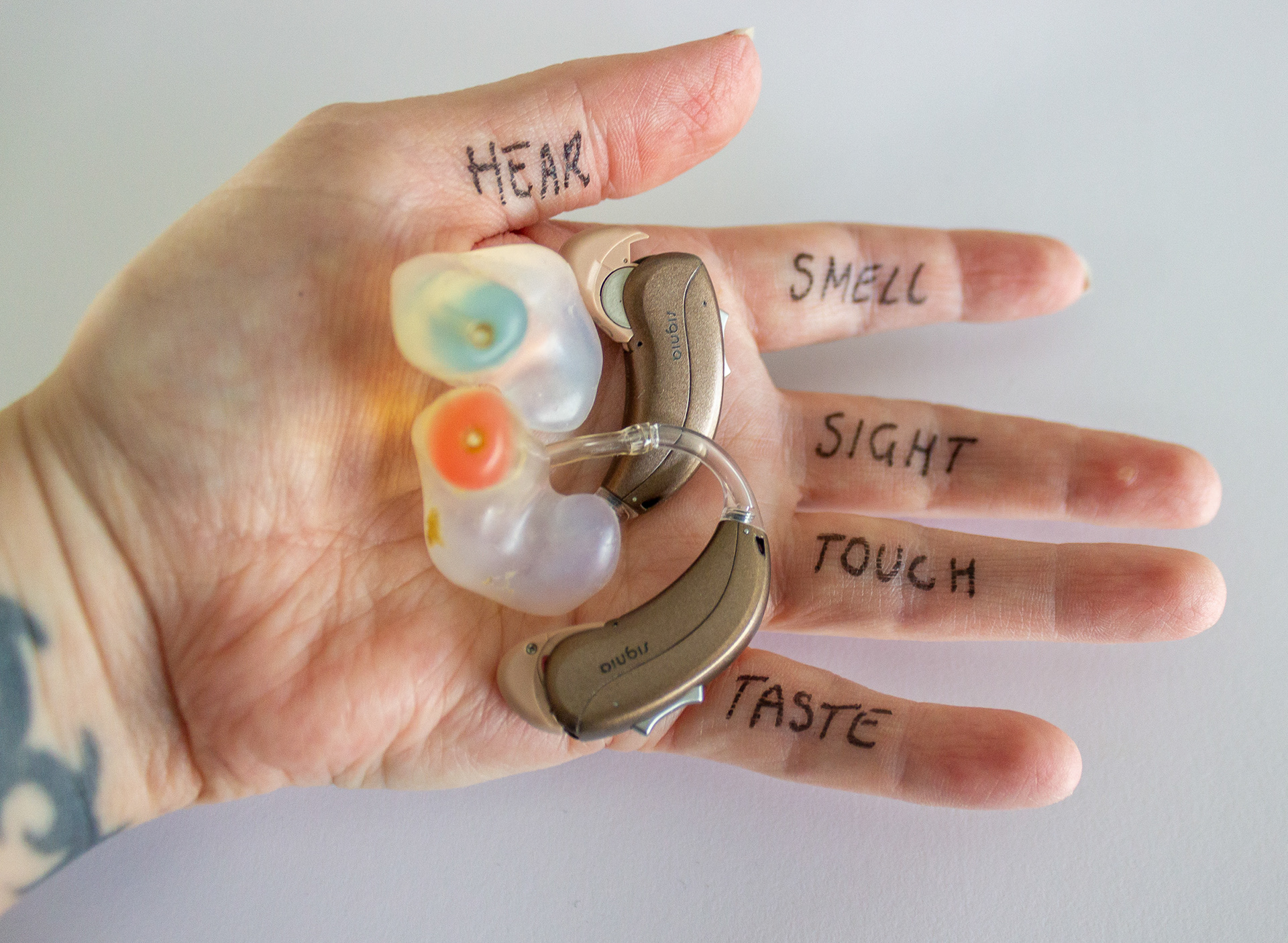 a close up on Mel's hand, with the words hear, smell, sight, touch, taste written on the hand. Also 2 hearing aids sit on her hand