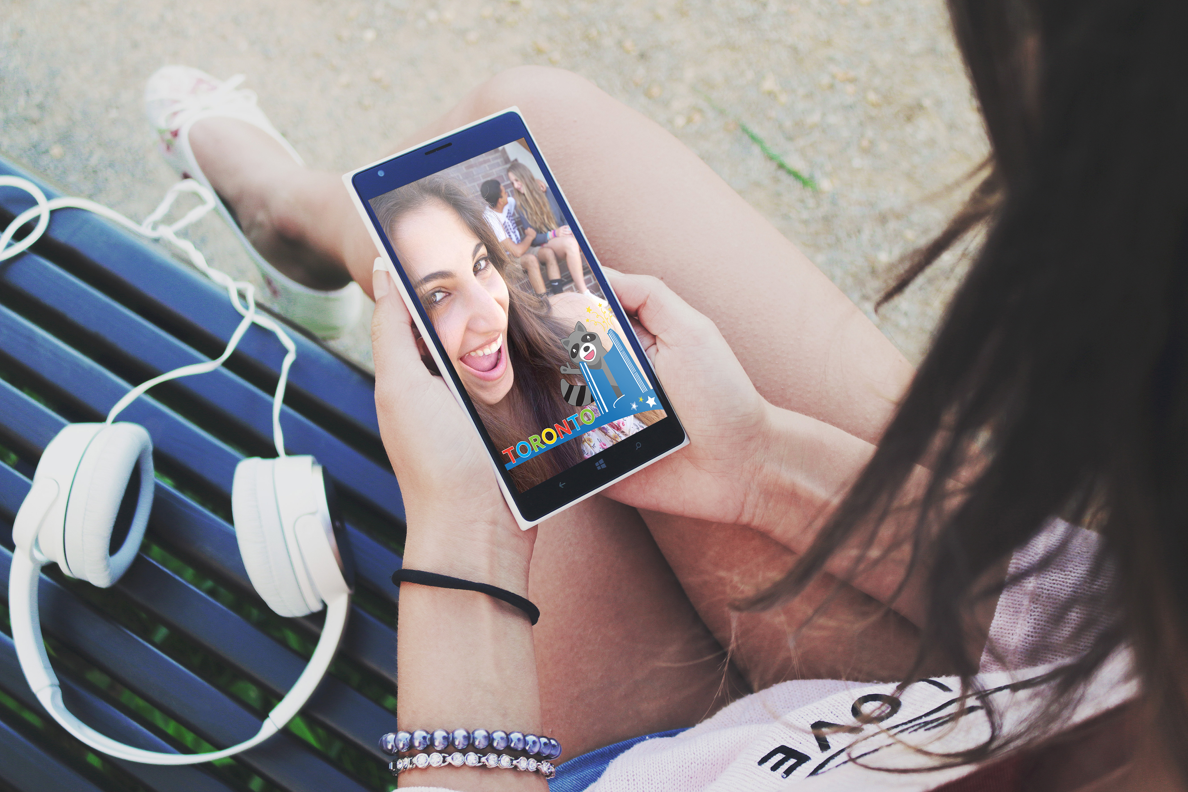 Photo of a teenage girl looking at her cell phone. On the screen is a smiling selfie with a Snapchat geofilter of a waving raccoon, City Hall, and the Toronto sign