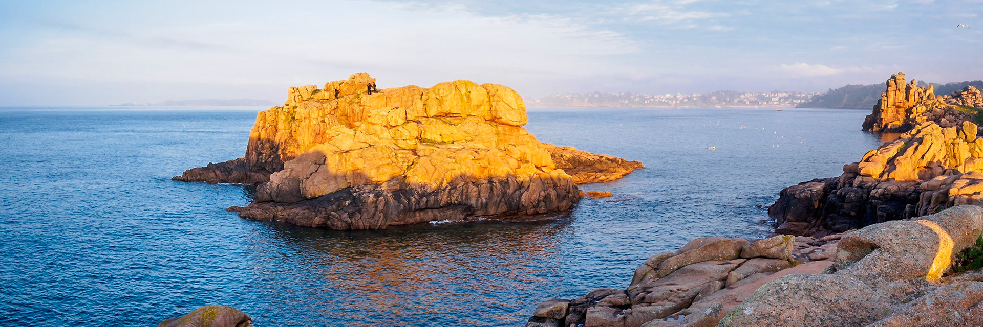 Under a blue sky during a golden sunset, a small group of daredevil divers prepared to jump off this cliff in Ploumanach, Bretagne. Wide shot with broad scenery of the pink granite, Perros-Guirec in the background.