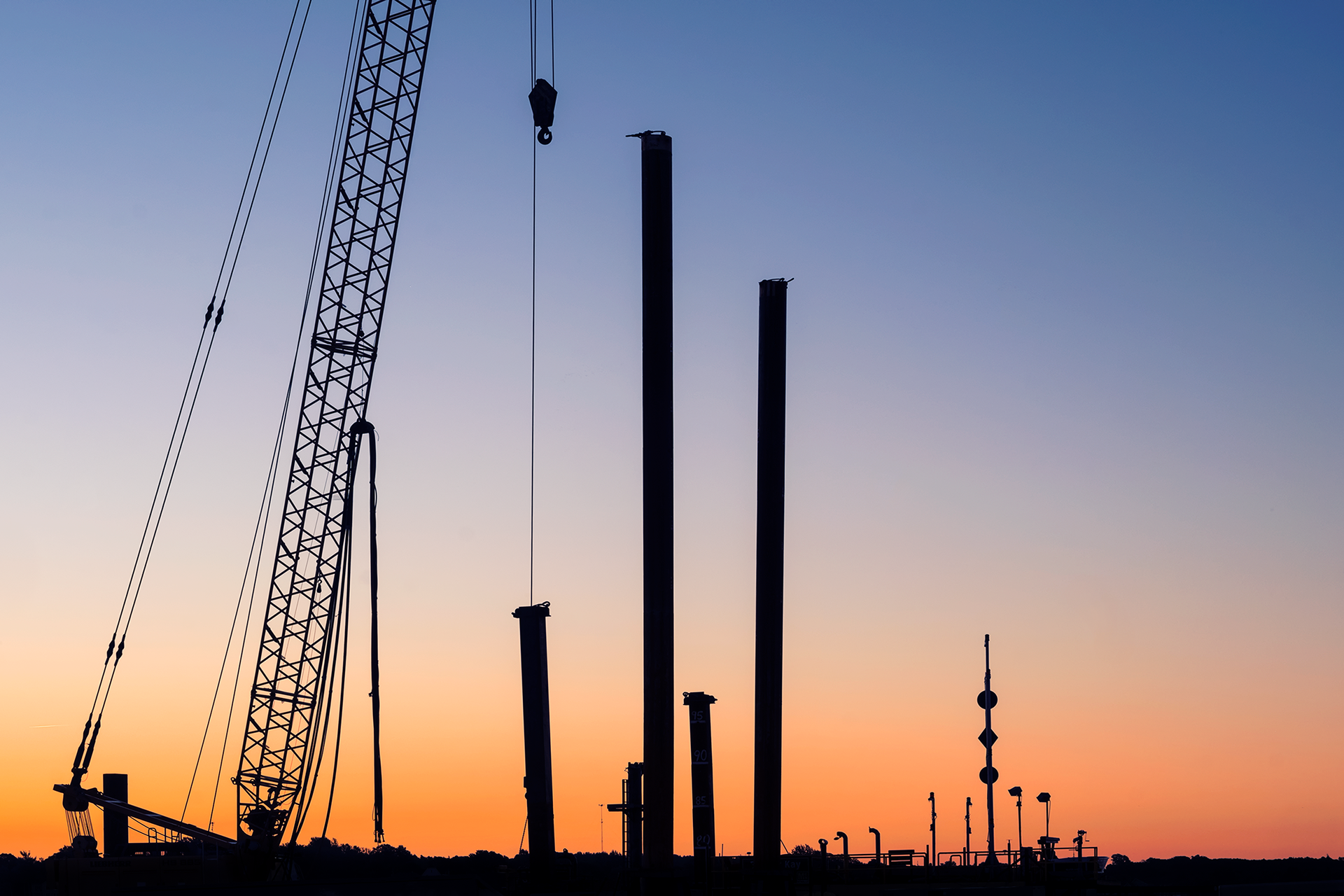 A crane dangles a pulley over four steel towers of a varying heights; the morning sky is gradient that runs from yellow to pale orange